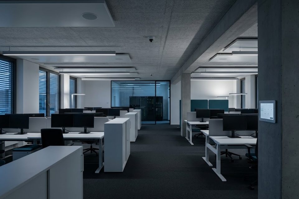 Bürogeschoss am Abend mit Arbeitsplätzen, Linienleuchten, Deckenlautsprechern, Kuppelkamera, Serverraum hinter Glas und Touchpanel an einer Säule.
